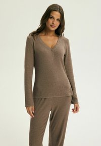 Brązowa bluzka z długim rękawem i dekoltem w kształcie litery V, z detalami w postaci guzików, w zestawie z dopasowanymi spodniami. Materiał jest teksturowany i miękki, co tworzy wygodny wygląd.