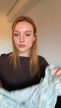 Jeune femme aux cheveux roux pliant un tissu clair à motifs à l'intérieur devant un mur uni.