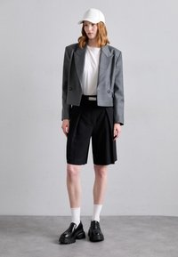 Sacou gri scurt peste un tricou alb, asociat cu pantaloni scurți pliți negri. Pantofi slip-on negri și șosete albe finalizează look-ul.