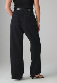 Črne široke kavbojke z visokim pasom, z zadnjimi žepi in gladko teksturo, kombinirane s črnimi sandali s peto.