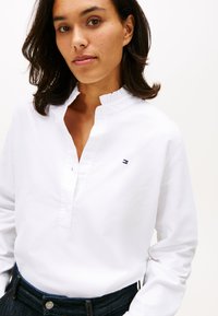 Žena s bílým tričkem s dlouhým rukávem a malým logem na hrudi, tmavými džínami a volnými tmavými vlasy přes ramena.