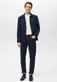 Marineblå knapstribet jakke med to brystlommer, parret med en hvid skjorte og mørke denimjeans, afsluttet med sorte sko.