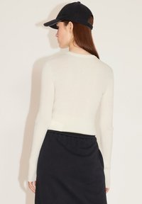 Weißes, geripptes Langarmoberteil, kombiniert mit einem schwarzen Rock. Das Model trägt eine schwarze Kappe; der Stoff ist weich und sitzt eng.
