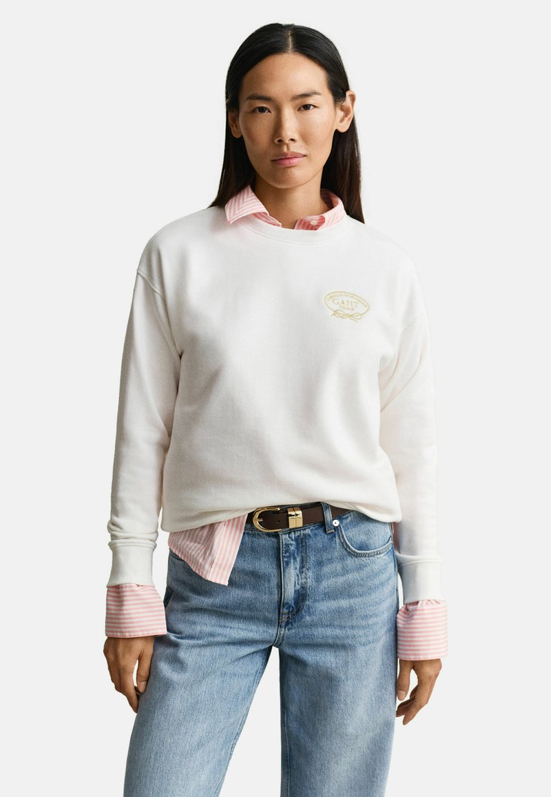 Femme portant un sweat-shirt blanc par-dessus une chemise rose à rayures rentrée dans un jean bleu avec une ceinture noire, debout devant un fond blanc.