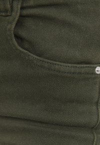 Gros plan sur un tissu en denim vert foncé montrant une poche avant avec des coutures visibles et un rivet argenté sur le bord.