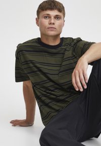 Camiseta de manga corta en color verde oliva con rayas verticales oscuras, cuello redondo acanalado en negro y corte holgado, combinada con pantalones negros.