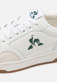 Sneaker in pelle bianca con logo verde, lacci bianchi e dettagli in suede beige. Suola in gomma solida con una superficie texturizzata.