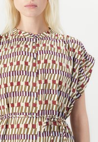 Knappet skjorte med en rund krave, der har et gentaget geometrisk mønster i dæmpede farver som oliven, maroon og creme. Bundet i taljen.