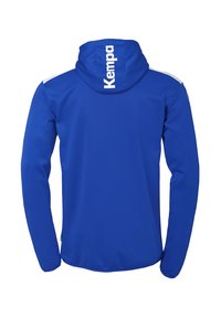 Blauwe sportjack met capuchon, lange mouwen en witte accenten. Heeft het 'Kempa' logo prominent op de achterkant en op de mouwen.