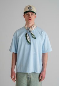 Light blue oversized t-shirt made from cotton, worn with a patterned silk scarf and beige cap featuring a green visor and logo.