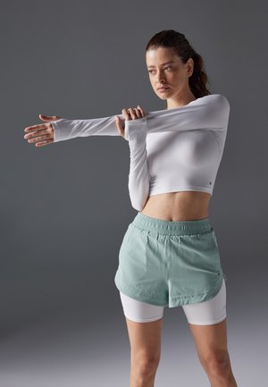 Hvit langt ermet cropped topp med tommelhull, kombinert med mintgrønne elastiske shorts over hvite tettsittende shorts, foran en grå bakgrunn.
