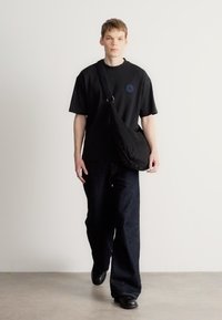 Μαύρο υπερμεγέθες μπλουζάκι με μπλε κυκλικό έμβλημα, συνδυασμένο με σκούρο χαλαρό τζιν και μαύρα παπούτσια, πάνω σε λευκό φόντο.