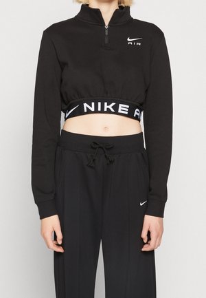 Sweatshirt noir court avec un col haut, doté d'une fermeture éclair à l'avant et du logo "Nike Air" blanc sur la taille. Associé à un pantalon de jogging noir.