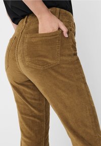 Pantalon en velours côtelé marron avec texture côtelée verticale, coupe taille mi-haute, et poches arrière, porté avec une main dans la poche droite.