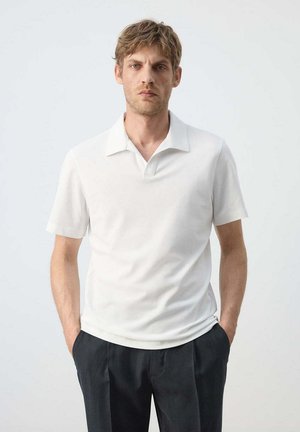 Homme aux cheveux courts châtain clair porte un polo blanc uni à manches courtes et un pantalon noir, debout les mains dans les poches devant un fond blanc.