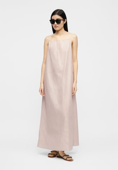 Ženska, ki nosi dolgo, svetlo rožnato obleko brez rokavov z ročnim vzorcem, črna sončna očala in rjave ravne sandale, stoji ob belem ozadju.