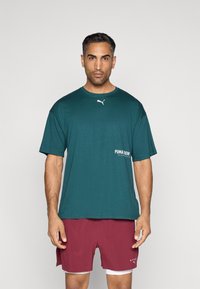 Zöld crew neck póló fehér Puma log�óval és szöveges részlettel elöl, bordó rövidnadrággal és fehér alsó réteggel párosítva.