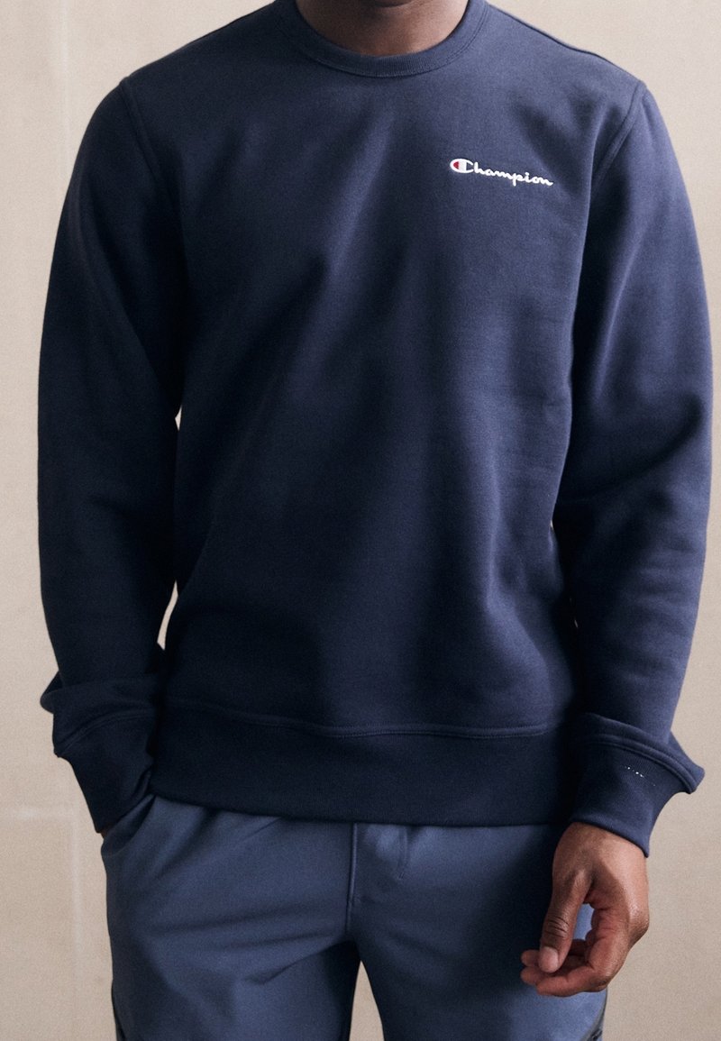 Personne portant un sweat-shirt Champion bleu marine avec les mains dans les poches sur un fond clair uni.