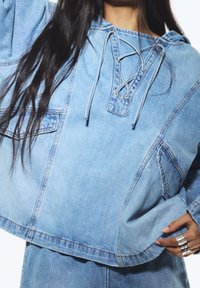 Felpa in denim di colore azzurro chiaro, con collo a imbuto e coulisse, vestibilità oversize e tasca frontale. Texture liscia con dettagli di cucitura a contrasto.