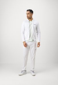 Ensemble de sport blanc comprenant une veste zippée et un pantalon assorti, avec un t-shirt vert clair en dessous et des accents de marque.