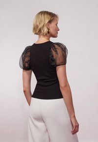 Black ribbed top with sheer puff sleeves, featuring a fitted silhouette. White wide-leg pants partially visible at the bottom.