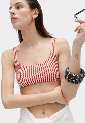Kobieta z mokrymi włosami, ubrana w czerwono-biały crop top w faliste paski, czarno-białe puszyste bransoletki, trzyma okulary przeciwsłoneczne, z tatuażem strzały na przedramieniu.