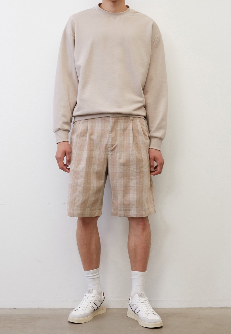 Sweatshirt beige à col rond, short à carreaux beige et marron clair, baskets blanches avec des accents noirs, chaussettes blanches, debout devant un fond uni.