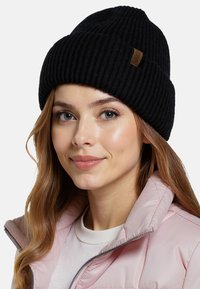 Czarna, prążkowana czapka typu beanie z podwiniętym mankietem i brązową skórzaną łatą. Noszona z jasnoróżową pikowaną kurtką.