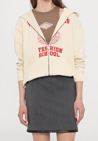 Donna che indossa una felpa beige con zip e testo rosso "Fashion School", un top senza maniche marrone con scollatura e una gonna grigia a coste con rifiniture nere.