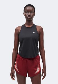 Jeune femme debout face à l’avant, portant un haut de sport noir sans manches et un short de course rouge avec des accents blancs et noirs.