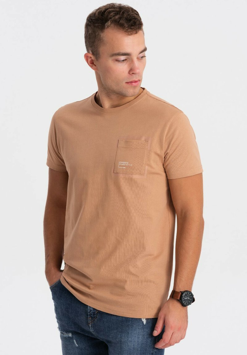 Tan kortærmet t-shirt med rund hals og detalje med lomme på venstre bryst. Sammen med slidte blå denimjeans og et mørkt ur.