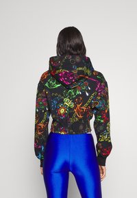 Giacca a cappuccio cropped in tessuto nero con motivi ornati multicolori. Presenta una vita e polsini elasticizzati, abbinata a leggings blu lucidi.