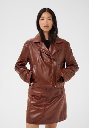 Femme aux longs cheveux foncés portant une veste en cuir marron avec fermeture éclair et une jupe en cuir marron assortie sur un fond uni.