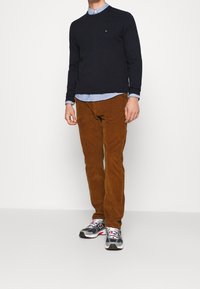 Maglione blu navy, camicia azzurra con bottoni sotto, pantaloni di velluto a coste marroni e sneakers grigie con accenti neri e rossi.