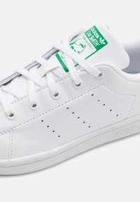 White leather sneaker with green accents and logo on the tongue, featuring round eyelets and perforated detailing on the sides.