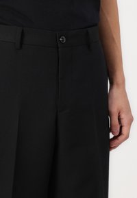 Pantalons noirs ajustés en tissu lisse, avec une taille plate, des poches latérales et une fermeture à bouton à la taille.