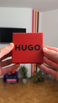Boîte carrée rouge avec le texte « HUGO » en lettres noires en gras sur le devant. Matériau lisse et brillant avec des bords légèrement arrondis.