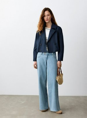 Jeune femme debout à l'intérieur, portant une veste courte bleu marine, un haut blanc, un jean clair à jambes larges, des chaussures beige et tenant un sac tissé à côté d'elle.