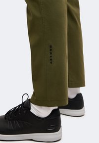 Jambes portant un pantalon vert olive avec le texte "Oakley" près de la cheville, des chaussettes blanches et des baskets noires avec des semelles blanches.