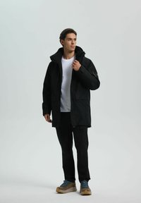 Giacca nera impermeabile con colletto alto, tasche frontali e polsini regolabili; indossata sopra una maglietta bianca e abbinata a pantaloni e scarpe nere.