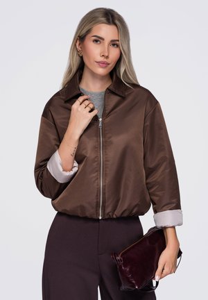 Femme aux longs cheveux blonds porte une veste marron zippée sur un haut gris, tenant une pochette marron foncé sur un fond uni.