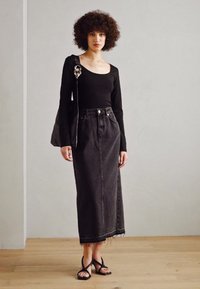 Top negro de manga larga combinado con una falda de mezclilla oscura de cintura alta, con dobladillo sin terminar. La modelo lleva sandalias negras con tiras. Fondo liso.