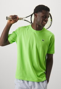 T-shirt di cotone verde neon con maniche corte, colletto a coste e un piccolo logo nero sul lato sinistro del petto. Il soggetto tiene in mano una racchetta da tennis.