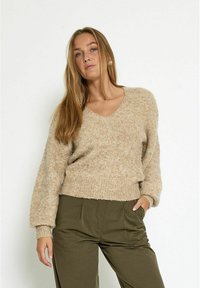 Beige V-hals trui met een zachte, gebreide textuur; heeft lange mouwen en een cropped, relaxte pasvorm. Combinatie met olijfgroene broek.