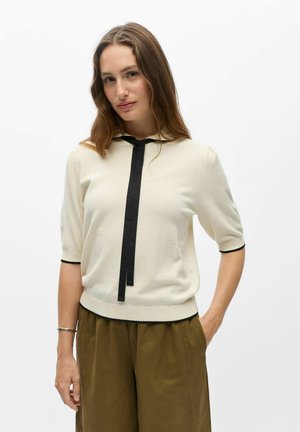 Femme aux longs cheveux bruns portant un pull crème à manches courtes avec un détail de nœud noir et un pantalon vert olive, debout avec une main dans la poche.