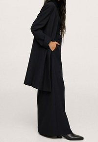 Zwarte op maat gemaakte lange jas met brede mouwen en zijzakken, gedragen over bijpassende donkere broek. Glad materiaal met een elegante afwerking.