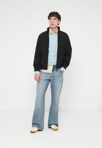 Μαύρο ελαφρύ μπουφάν πάνω από ένα γαλάζιο πουλόβερ, σε συνδυασμό με πουκάμισο με γιακά και ξεβαμμένο τζιν καμπάνα σε μπλε απόχρωση. Το λευκά αθλητικά παπούτσια ολοκληρώνουν το σύνολο.