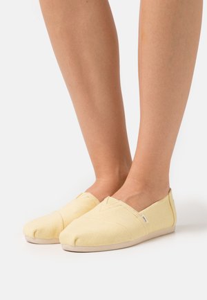 Yellow canvas slip-on shoes with closed toes, stitched detailing, and a light-colored sole. Designed for comfort with soft texture.