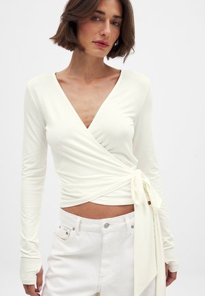 Romantische crèmekleurige longsleeve wikkeltop met een diepe V-hals en een zijstrikdetail, gemaakt van zacht materiaal, gecombineerd met witte high-waisted jeans.