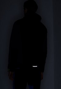 Personne vue de dos portant une veste sombre avec une petite bande réfléchissante près du bas du dos, se tenant devant un fond sombre.